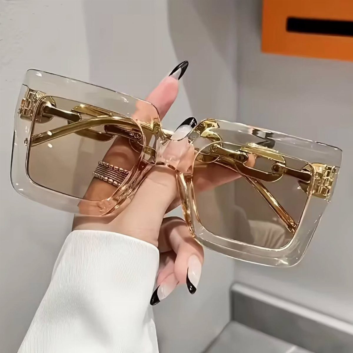 Oversized square sunglasses with transparent frames and gold chain details held by a hand.