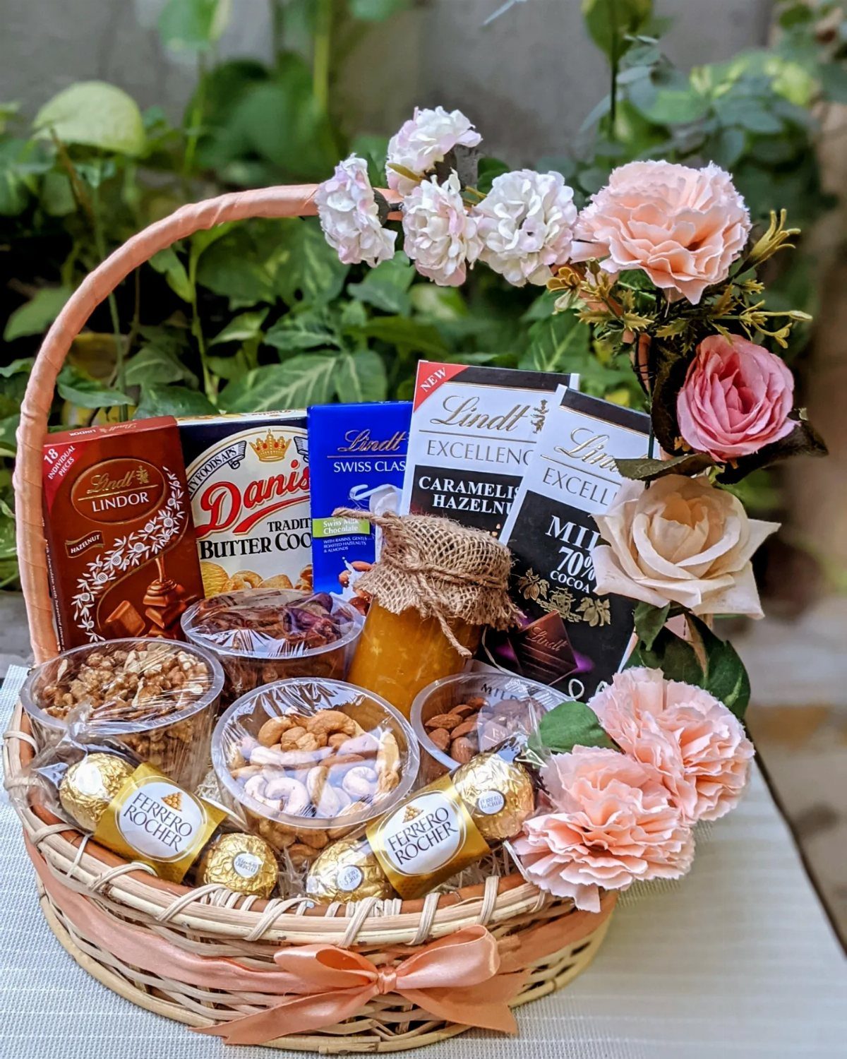 Wicker gift basket filled with chocolates, honey, and nuts, decorated with pink roses.