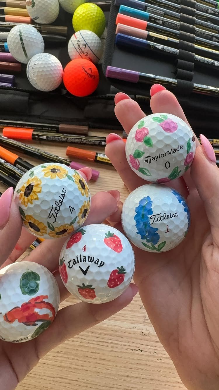 Hands holding white golf balls painted with colorful flowers and strawberries.