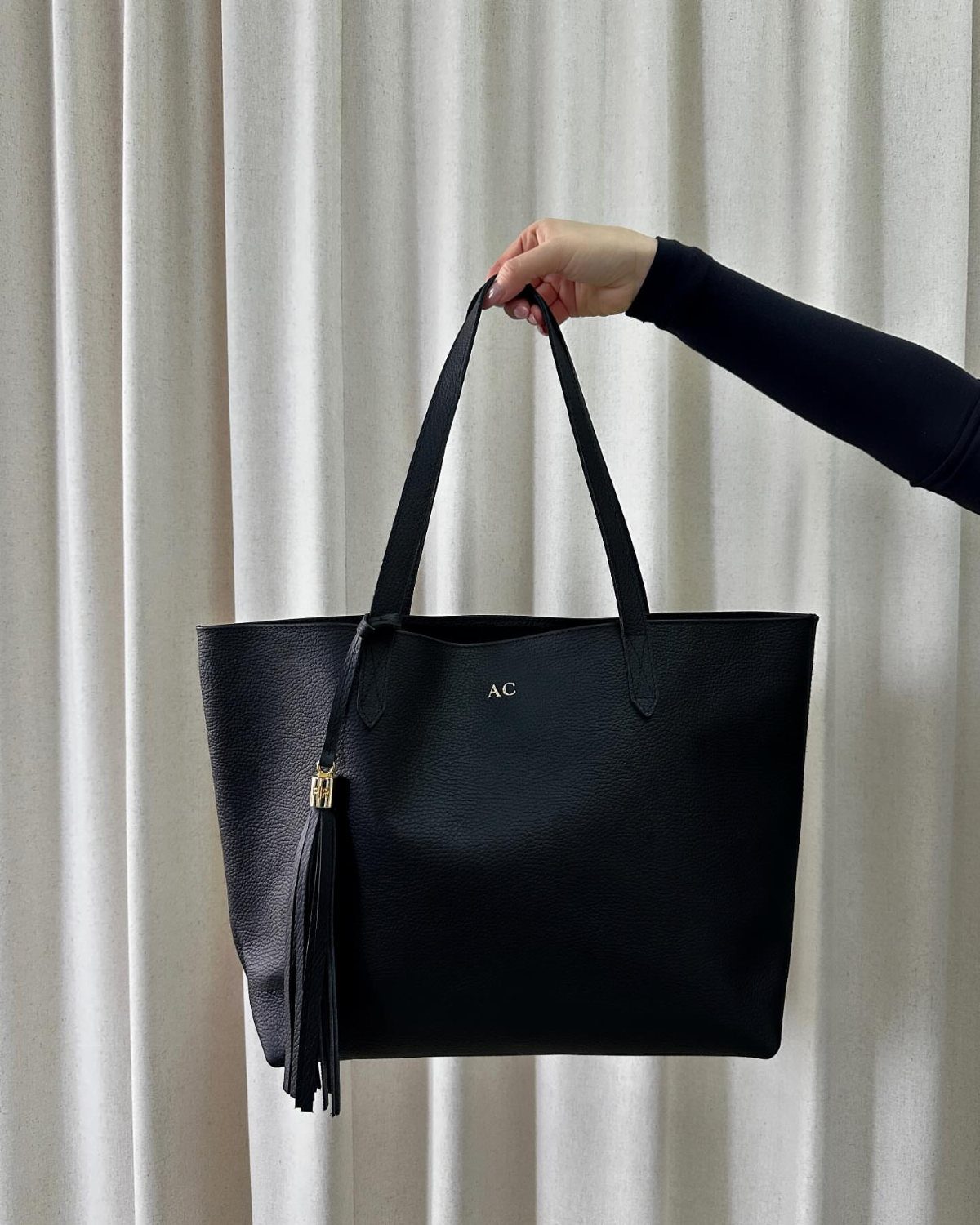 Black leather tote bag with gold personalized initials held against white curtains