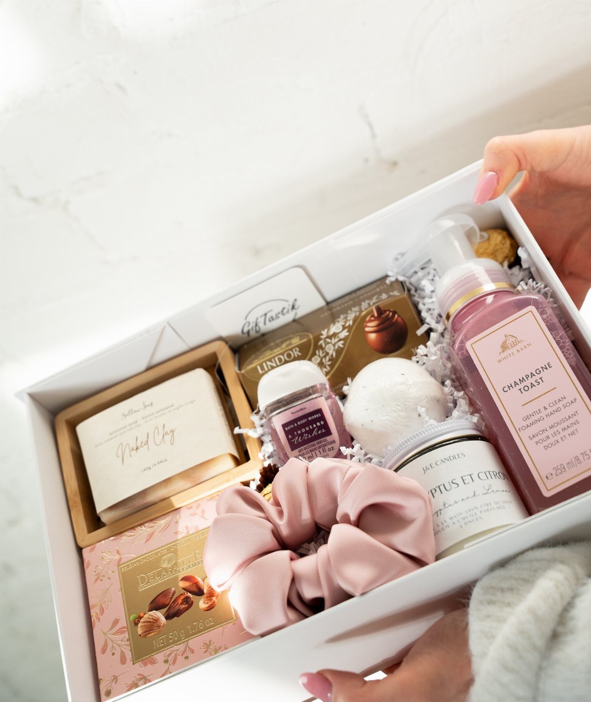 Gift box with pink soap, bath bomb, scrunchie, and chocolates on white filler.
