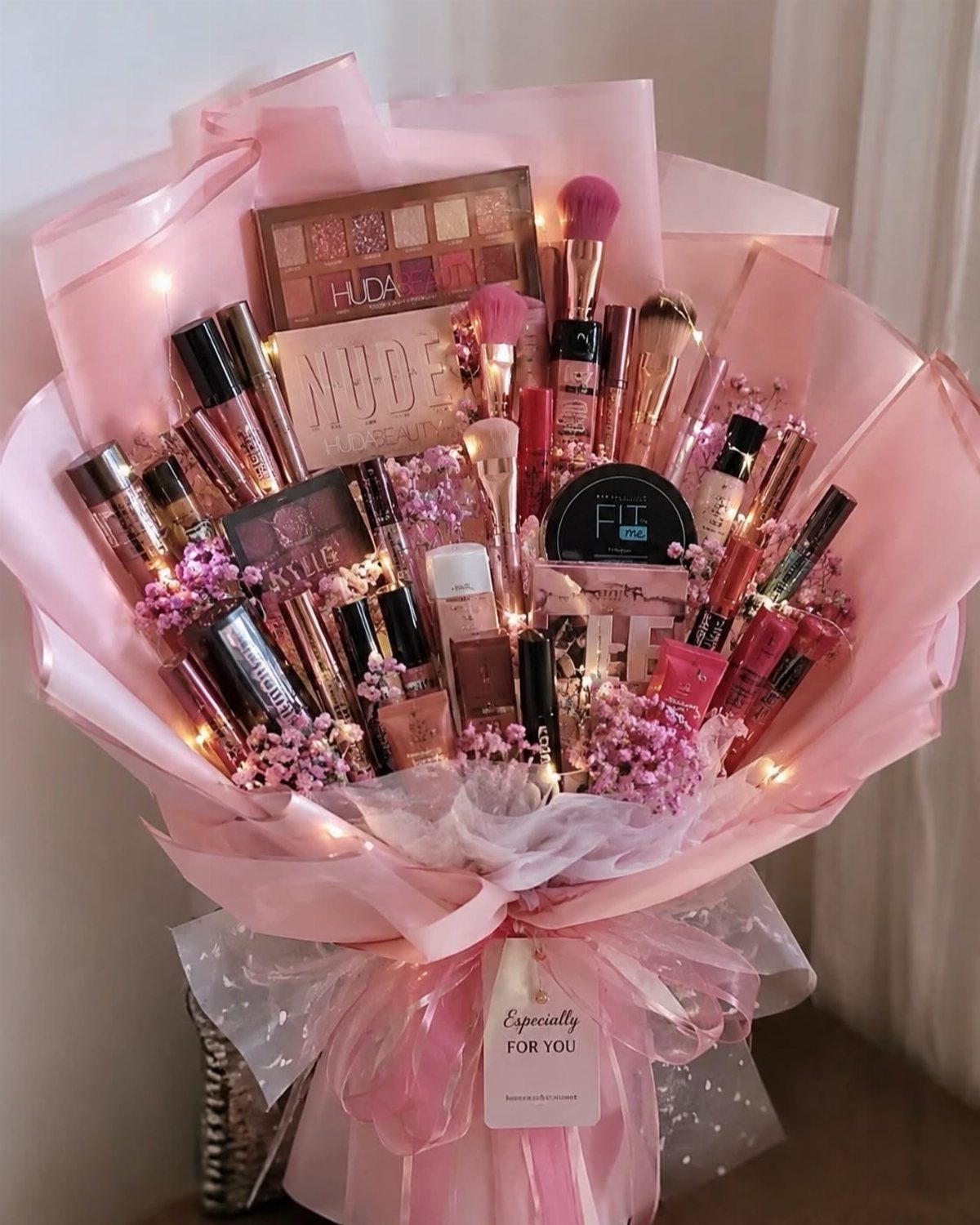 Makeup bouquet with cosmetics, pink flowers, and fairy lights wrapped in pink paper.