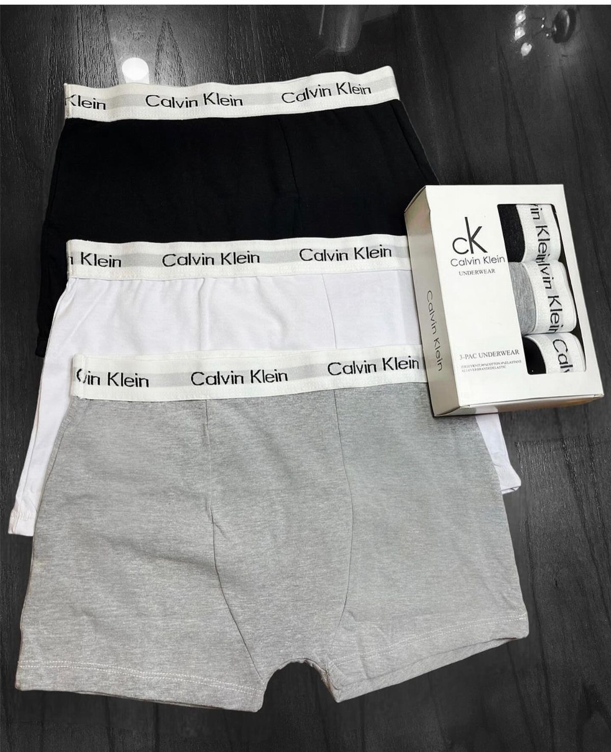 Three Calvin Klein boxer briefs in black, white, and grey with branded packaging.