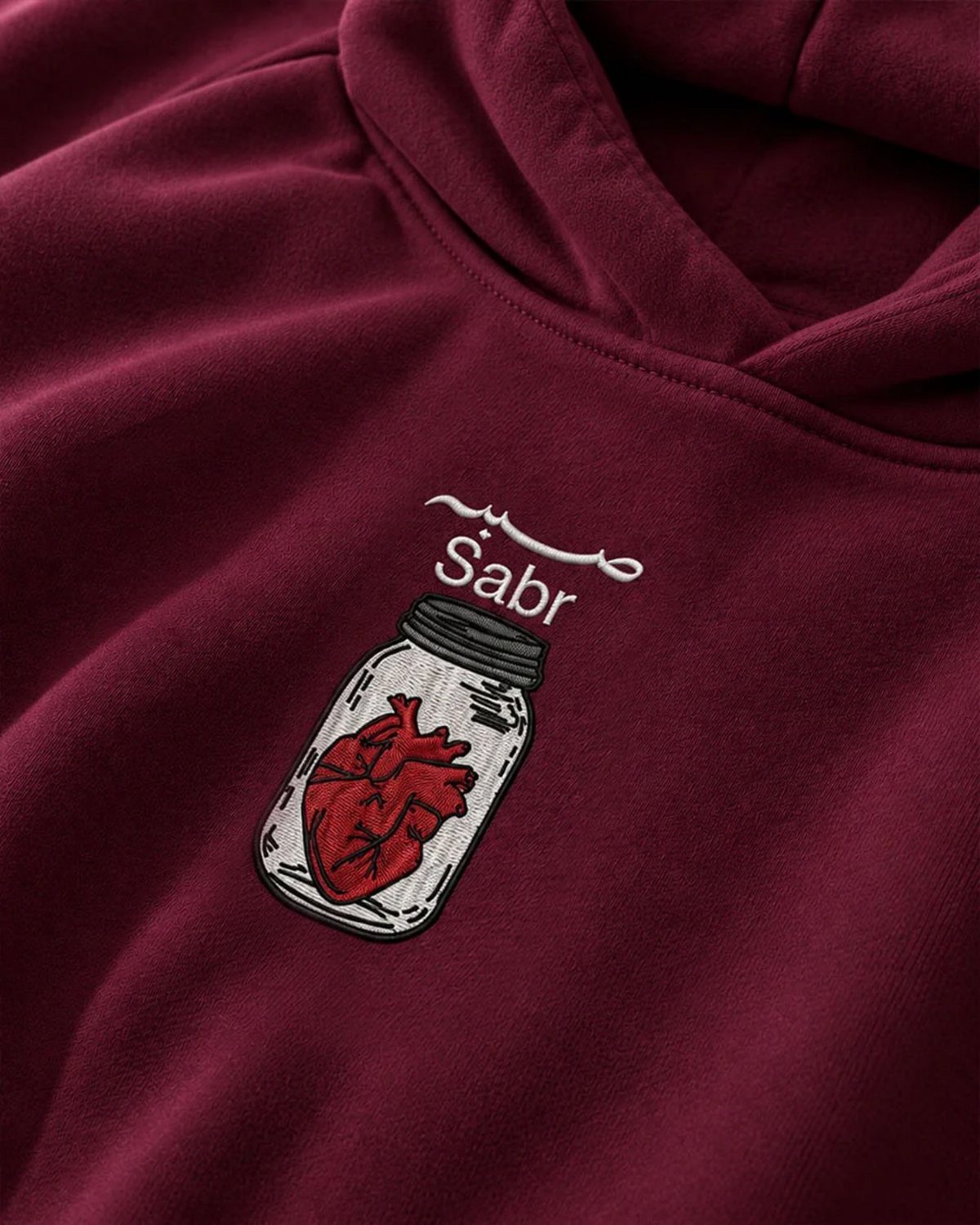 Maroon hoodie with an embroidered heart in a jar and the word Sabr.