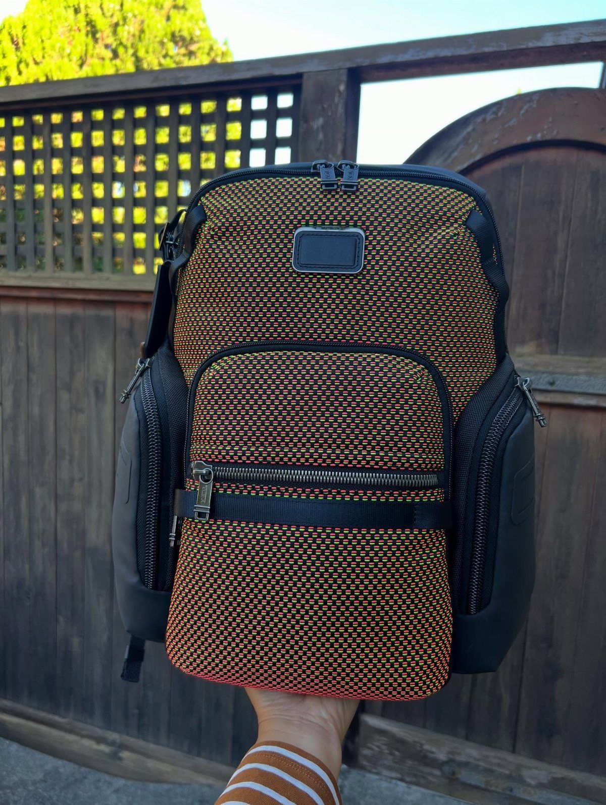 Woven orange and black backpack held in front of a wooden fence.