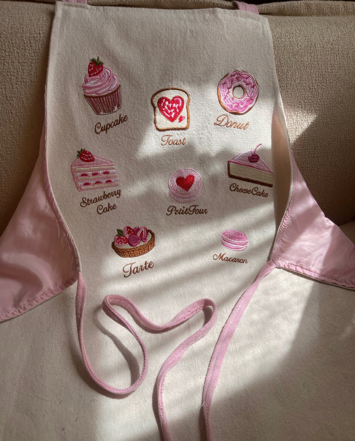 Cream apron with pink trim and embroidered dessert illustrations and labels.