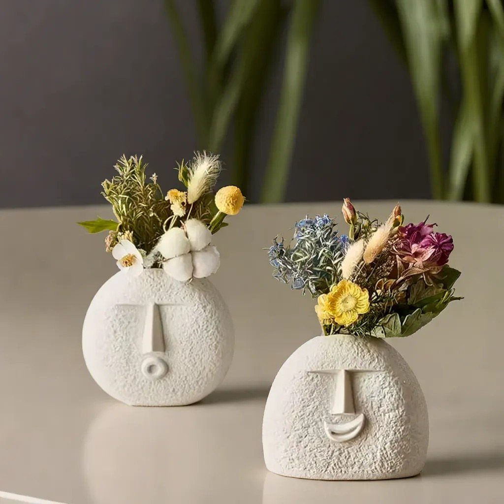 Two white face-shaped ceramic vases holding colorful dried flower arrangements on a table.