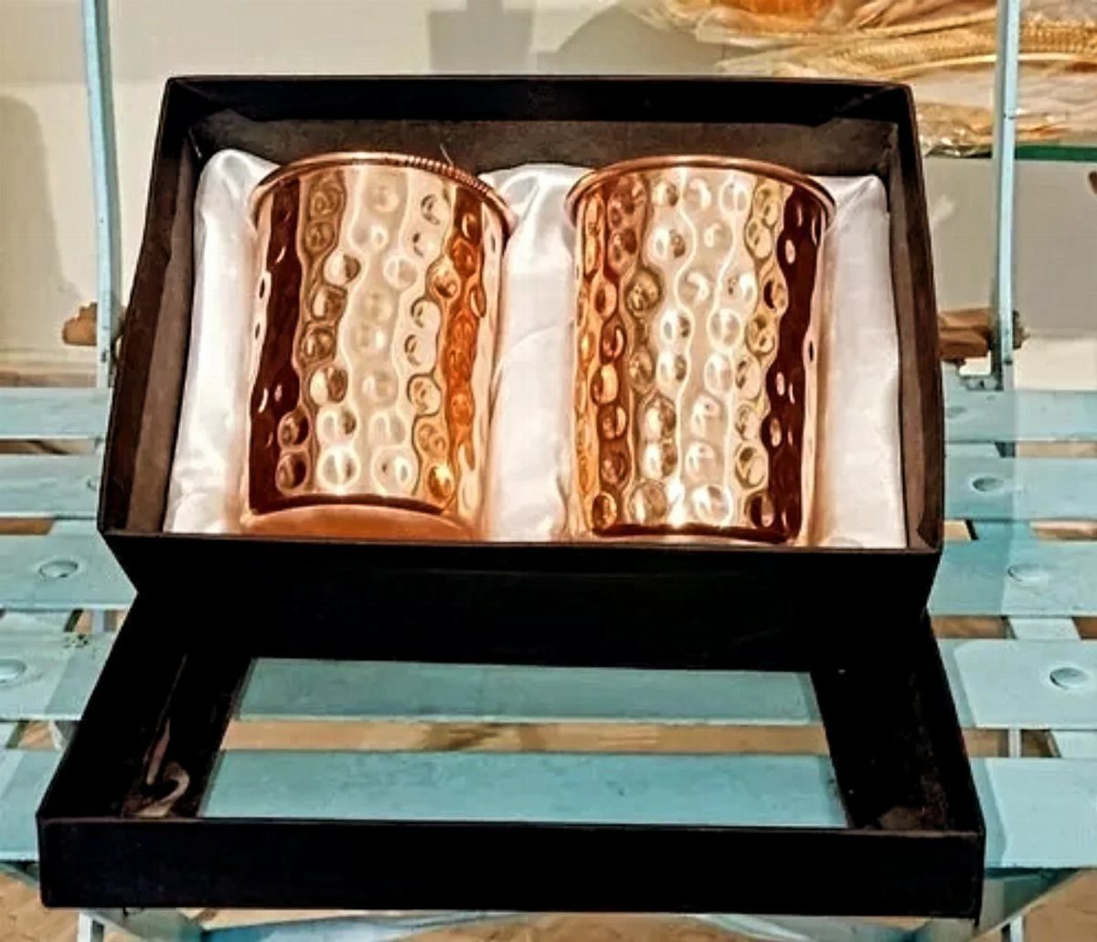 Two hammered copper mugs in a black gift box on a blue surface.