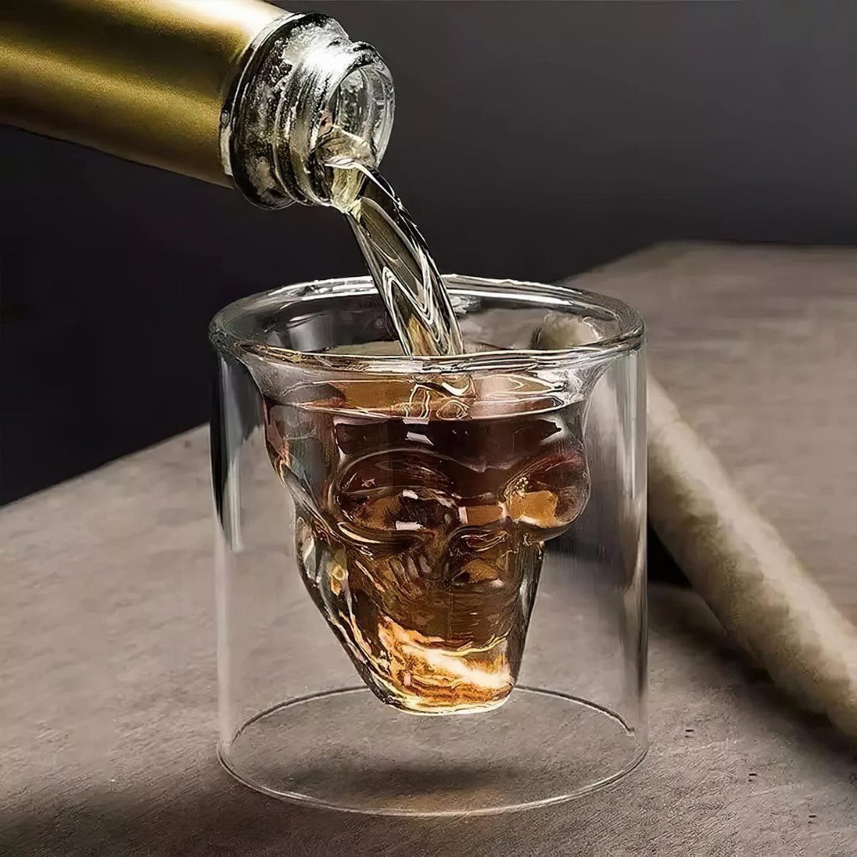 Amber liquid pouring from a gold bottle into a skull-shaped double-walled glass.