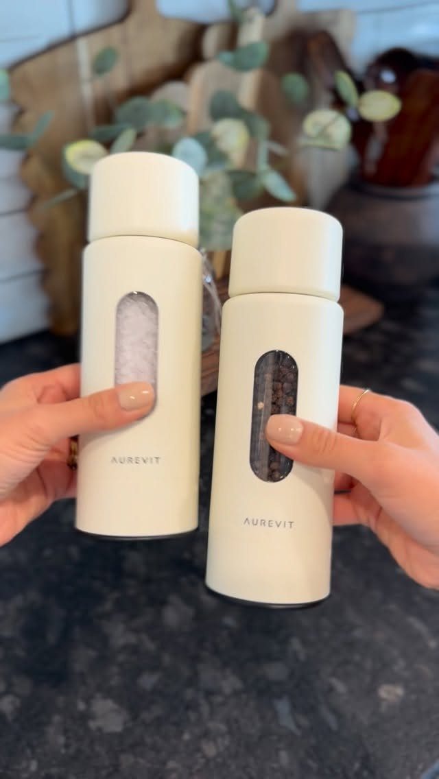 White electric salt and pepper grinders held by hands in a kitchen setting.