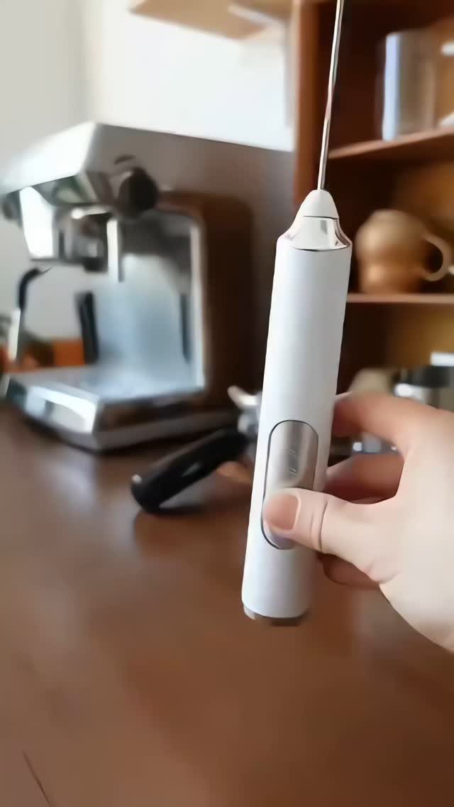 White handheld milk frother held by a hand with an espresso machine background.
