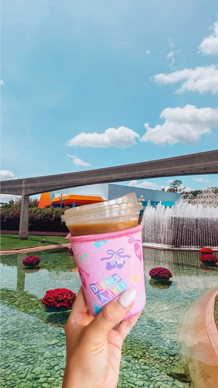 Iced coffee in a pink insulated sleeve held against a sunny outdoor fountain background