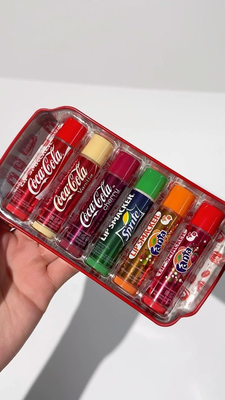 Six soda-themed lip gloss tubes in a red plastic tray held by a hand.